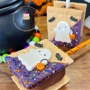 May include: Two graham cracker s'mores decorated with purple and white icing, sprinkles, and a ghost-shaped marshmallow. The s'mores are on a wooden surface with a black cauldron and a jack-o-lantern in the background.