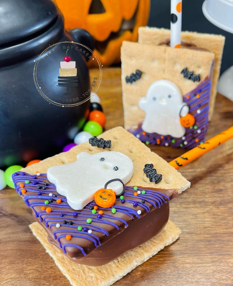 May include: Two graham cracker s'mores decorated with purple and white icing, sprinkles, and a ghost-shaped marshmallow. The s'mores are on a wooden surface with a black cauldron and a jack-o-lantern in the background.