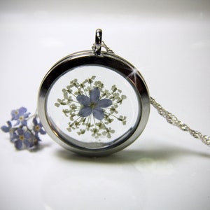 May include: A silver locket necklace with a clear glass insert containing a pressed blue flower and white flower buds. The chain is silver and has a delicate design.