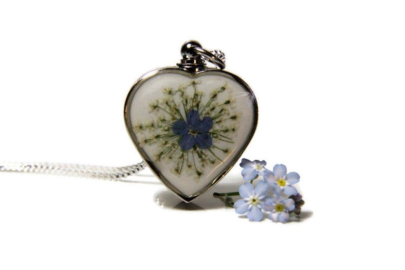 K&ouml;nnte beinhalten: Eine silberne herzf&ouml;rmige Halskette mit einem gepressten Vergissmeinnicht-Blumenanh&auml;nger. Die Blume ist blau mit einer wei&szlig;en Mitte und von gr&uuml;nen Bl&auml;ttern umgeben.
