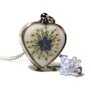 K&ouml;nnte beinhalten: Eine silberne herzf&ouml;rmige Halskette mit einem gepressten Vergissmeinnicht-Blumenmotiv. Der Anh&auml;nger ist an einer silbernen Kette und es liegen Vergissmeinnicht-Blumen auf dem Tisch darunter.