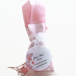 May include: A pink and white gift bag with a white flower and pearl embellishment. The bag has a round white tag with the text "Jasmine Thyme Designs" and website information.