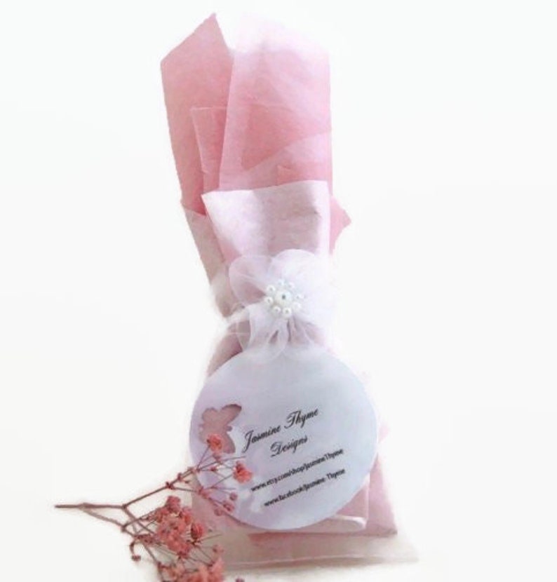 May include: A pink and white gift bag with a white flower and pearl embellishment. The bag has a round white tag with the text "Jasmine Thyme Designs" and website information.