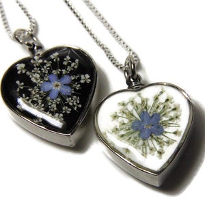 K&ouml;nnte beinhalten: Zwei silberne herzf&ouml;rmige Halsketten mit einer blauen Blume und wei&szlig;en Blumen, die in das Glas gepresst sind. Die Halsketten liegen auf einem wei&szlig;en Hintergrund.