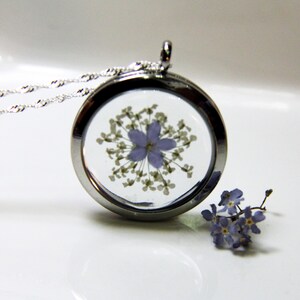 May include: A silver locket necklace with a delicate chain. The locket contains a pressed purple flower with white petals. A small cluster of purple flowers is visible in the background.