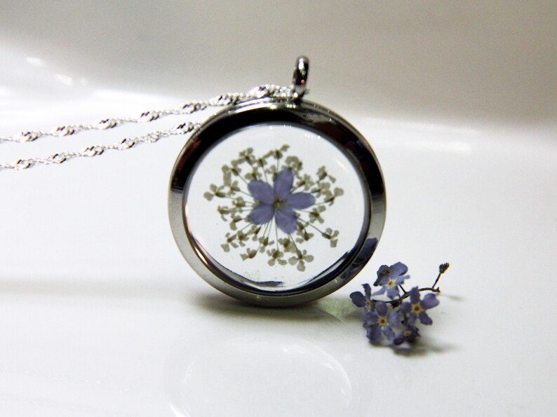 May include: A silver locket necklace with a delicate chain. The locket contains a pressed purple flower with white petals. A small cluster of purple flowers is visible in the background.