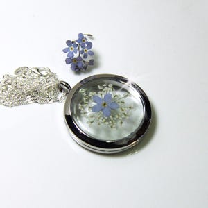 May include: A silver locket necklace with a clear glass insert containing a pressed blue flower and white flowers. The necklace has a silver chain.