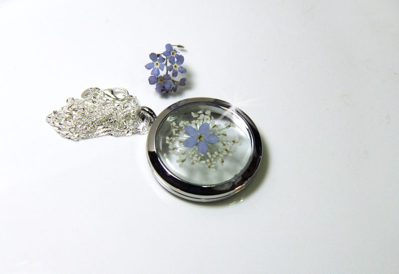 May include: A silver locket necklace with a clear glass insert containing a pressed blue flower and white flowers. The necklace has a silver chain.