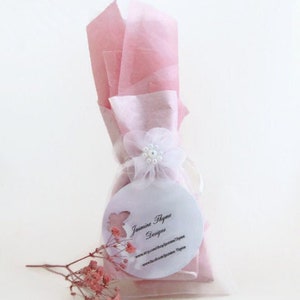 K&ouml;nnte beinhalten: Eine rosa und wei&szlig;e Geschenkt&uuml;te mit einer wei&szlig;en Blume und einem wei&szlig;en runden Etikett mit dem Text "Jasmine Thyme Designs" und der Website-Adresse "www.etsy.com/shop/JasmineThyme" und "www.facebook.com/Jasmine.Thyme".
