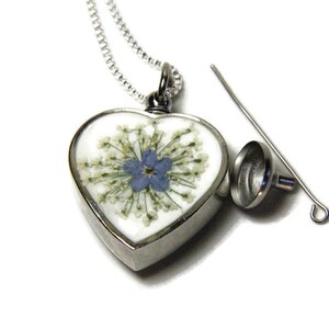 K&ouml;nnte beinhalten: Eine silberne herzf&ouml;rmige Halskette mit einem kleinen blauen Blumen- und wei&szlig;en Blumen-Druck im Inneren. Der Anh&auml;nger l&auml;sst sich &ouml;ffnen und zeigt ein kleines Fach zum Aufbewahren von Asche oder anderen Erinnerungsst&uuml;cken. Er wird mit einem kleinen Trichter und einer Kette geliefert.