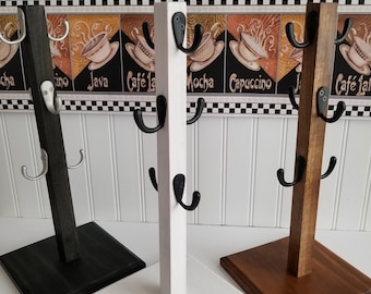 May include: Three wooden mug racks in black, white, and brown. Each rack has a square base and a vertical post with three black hooks for hanging mugs. The background features a coffee-themed border.