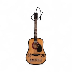 May include: Acoustic guitar ornament with a light brown body, black trim, and a black pickguard. The guitar features the text "NASHVILLE" and the number "25". The headstock has a black loop and a signature.