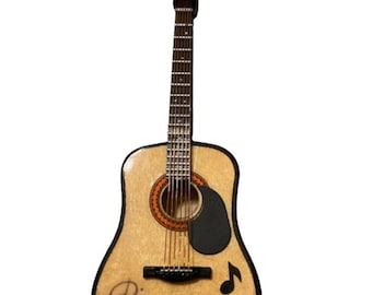Pode incluir: Miniatura de enfeite de guitarra acústica com acabamento em madeira clara, pickguard preto e o nome "REBA" impresso no corpo. Apresenta uma corda preta para pendurar. Inclui uma nota musical e uma assinatura.