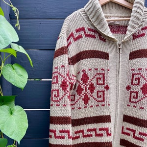 May include: A cream-coloured knitted cardigan with a brown and red geometric pattern. The cardigan has a zip closure and a collar.