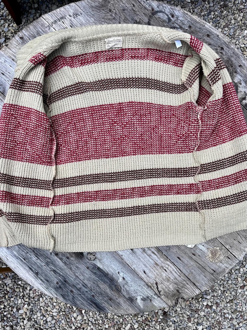 May include: A beige striped cardigan with red and brown stripes. The cardigan is unbuttoned and laid flat on a wooden surface.