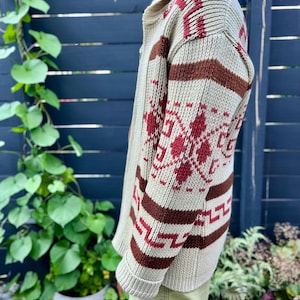 May include: A cream-coloured cardigan with a brown and red geometric pattern. The cardigan has a zip closure and a collar.