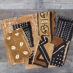 May include: A collection of brown, black, and tan fabric pieces with various geometric and abstract patterns. The textiles feature designs in white and tan, with some pieces displaying repeating shapes and symbols. The fabrics are arranged on a wooden surface.