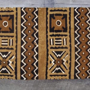 Può includere: Tessuto rettangolare con un motivo ripetuto di forme geometriche in marrone, nero e bianco. Il design include quadrati, zigzag ed elementi lineari. Il tessuto ha un aspetto strutturato e un bordo sfrangiato.