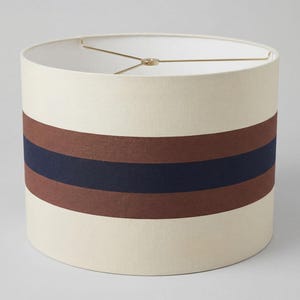 May include: A cylindrical drum lampshade featuring a cream-colored fabric exterior with a wide horizontal band consisting of a central dark navy blue stripe flanked by two brown stripes. The interior includes a gold-toned metal spider fitter.