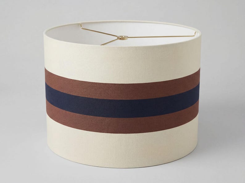 May include: A cylindrical drum lampshade featuring a cream-colored fabric exterior with a wide horizontal band consisting of a central dark navy blue stripe flanked by two brown stripes. The interior includes a gold-toned metal spider fitter.