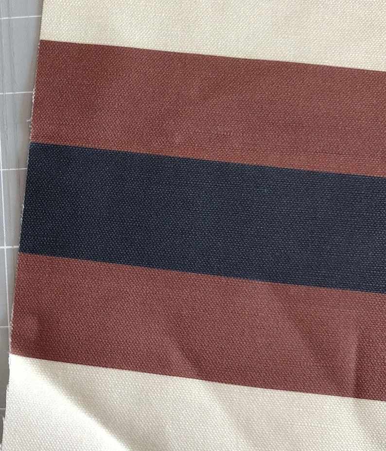 May include: A woven fabric featuring a repeating horizontal striped pattern. The design consists of wide bands in cream, followed by a thick stripe of dark brown, a central stripe of dark navy blue, and another thick stripe of dark brown.