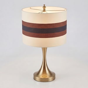 May include: A table lamp with a brushed brass finish base and a cylindrical drum shade. The shade is cream-colored with two horizontal bands in dark brown and black encircling the center. A small brass finial sits atop the shade frame.