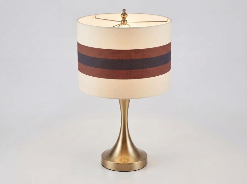 May include: A table lamp with a brushed brass finish base and a cylindrical drum shade. The shade is cream-colored with two horizontal bands in dark brown and black encircling the center. A small brass finial sits atop the shade frame.