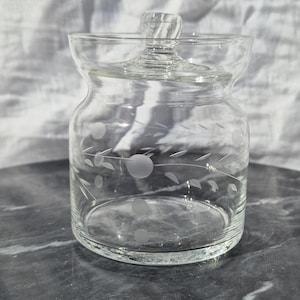 May include: A clear glass jar with a lid, featuring an etched design of leaves and dots. The jar is sitting on a dark grey surface.