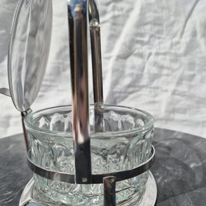 May include: A silver sugar bowl with a glass bowl and a hinged lid. The bowl has a decorative pattern and sits on a silver base.