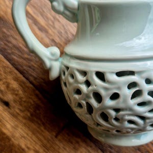 May include: A light green ceramic creamer with an intricate cut-out design. The creamer has a handle and a rounded body.