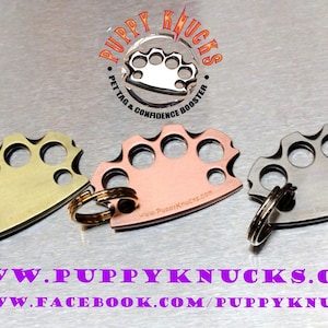 May include: Three metal keychains shaped like brass knuckles. The keychains are gold, copper, and silver. The keychains have the text "Puppy Knucks" and "Pet Tag & Confidence Booster" on them. The keychains are on a silver background.