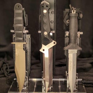 Puede incluir: Tres cuchillos tácticos exhibidos en un soporte de acrílico transparente. Los cuchillos tienen mangos negros y hojas plateadas. El cuchillo central tiene un diseño único. Las hojas son afiladas y puntiagudas, y los cuchillos están en sus fundas.