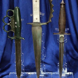 May include: Three vintage knives displayed on a clear acrylic stand. The knives include a green brass knuckle knife, a large knife with a handle guard, and a knife with a ribbed handle. The blades are various lengths and shapes.