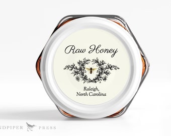 May include: A white jar with a hexagonal lid, filled with honey. The jar has a white label with black text that reads "Raw Honey" and a black and white illustration of a bee. The label also includes the text "Raleigh, North Carolina".