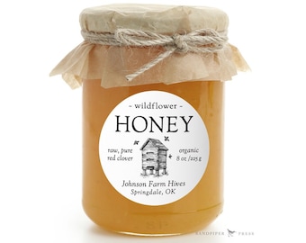 May include: A round jar of honey with a white label that says "wildflower - HONEY" and includes details about the honey, such as "raw, pure red clover" and "organic 8 oz/225 g". The label also includes the name of the producer, "Johnson Farm Hives", and their location, "Springdale, OK".