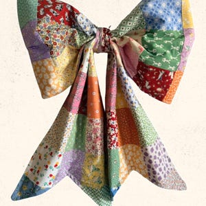 May include: A large, colorful patchwork bow with a variety of floral and geometric patterns. The bow is made of fabric squares in shades of red, blue, yellow, and green. The text "Sweet Lorraine Co." is at the bottom.