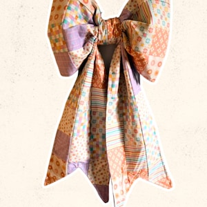May include: A large, colorful bow made of patchwork fabric. The bow features a variety of patterns, including floral, striped, and checkered designs in shades of orange, pink, purple, and yellow. The text "Sweet Lorraine Co." is written in cursive at the bottom.