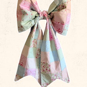 May include: A large, decorative bow made from patchwork fabric in pastel pink, blue, and green hues. The bow features a floral pattern and the text "Sweet Lorraine Co." in cursive script.