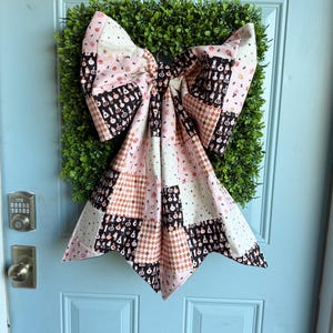 May include: A large fabric bow featuring a patchwork design with alternating squares of black, white, and soft pink. The pattern includes illustrations of small potion bottles, diamonds, and stars. The bow is mounted over a square green boxwood wreath.