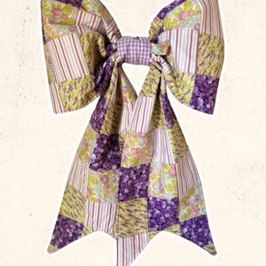 May include: A large, decorative bow featuring a patchwork design in shades of purple, lavender, and green. The bow is made of various patterned fabrics, including floral and striped patterns. The text "Sweet Lorraine Co." is written in cursive at the bottom.