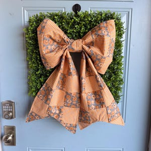 May include: A square green boxwood wreath adorned with a large fabric bow. The bow features a patchwork design alternating between a gray fabric with a repeating orange pumpkin pattern and an orange and white gingham checkered fabric pattern.