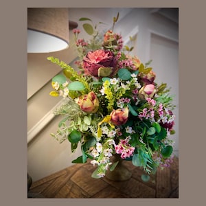 May include: A floral arrangement featuring a variety of flowers in shades of pink, red, and yellow, with green foliage. The bouquet is in a bronze-colored vase, placed on a wooden surface. The lighting is soft, creating a warm ambiance.