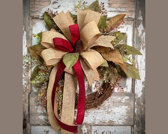 May include: A rustic wreath with a woven base, adorned with burlap and burgundy ribbons, and faux leaves. The wreath features a large bow and a pinecone in the center, creating a warm, inviting aesthetic. The wreath is displayed against a distressed, white-painted wooden door.