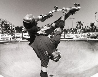 May include: A skateboarder performs a trick in a halfpipe, upside down with their legs extended and the skateboard above their head. The skateboarder is wearing protective gear, including knee pads and a helmet. The halfpipe is made of concrete and has a smooth surface. The background shows a crowd of people watching the skateboarder.