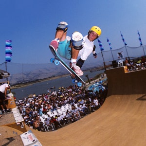 May include: A skateboarder wearing a yellow helmet and blue shorts performs a trick in a halfpipe. The halfpipe is made of wood and has a white concrete lip. The skateboarder is in mid-air, with his board facing the camera. There are many people watching the skateboarder in the background.