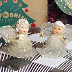 May include: Two vintage Christmas angel figurines. Each angel has a porcelain face, white hair, and sheer wings. One angel holds a star-topped wand. They are dressed in cream-colored gowns with silver glitter accents. The background includes a Christmas tree illustration.