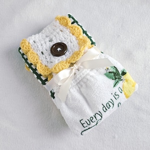 May include: A white crochet towel holder with a yellow scalloped edge and a brown button closure. The towel has a white ribbon and a printed lemon design with the text "Every day is a".