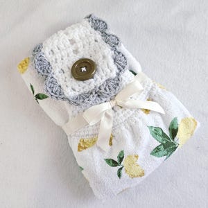 May include: A white hand towel with a lemon and leaf pattern, wrapped and tied with a cream-colored ribbon. The top features a white crocheted square with a gray scalloped edge and a brown button.