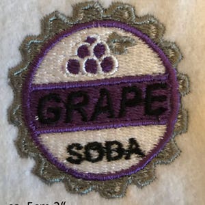 Puede incluir: Un parche bordado de color morado y blanco con un dise&ntilde;o de uva y las palabras "GRAPE SODA" en letras negras. El parche tiene un di&aacute;metro de aproximadamente 5 cm.