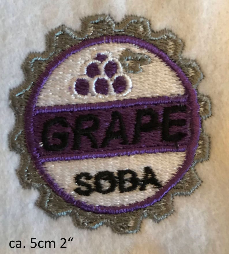 Puede incluir: Un parche bordado de color morado y blanco con un dise&ntilde;o de uva y las palabras "GRAPE SODA" en letras negras. El parche tiene un di&aacute;metro de aproximadamente 5 cm.
